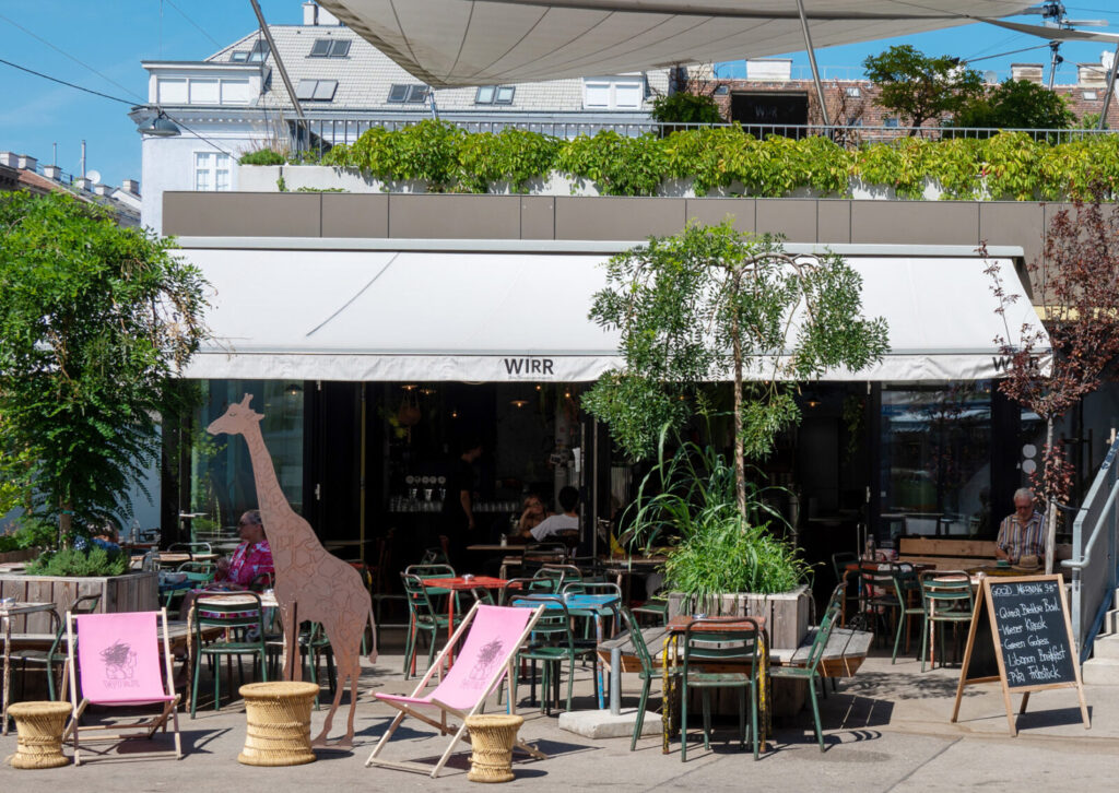 Außenbereich eines Cafés Wirr am Yppenplatz mit Tischen, Stühlen, Liegestühlen, einer großen Girafenskulptur und einer Tafel mit handgeschriebenem Text